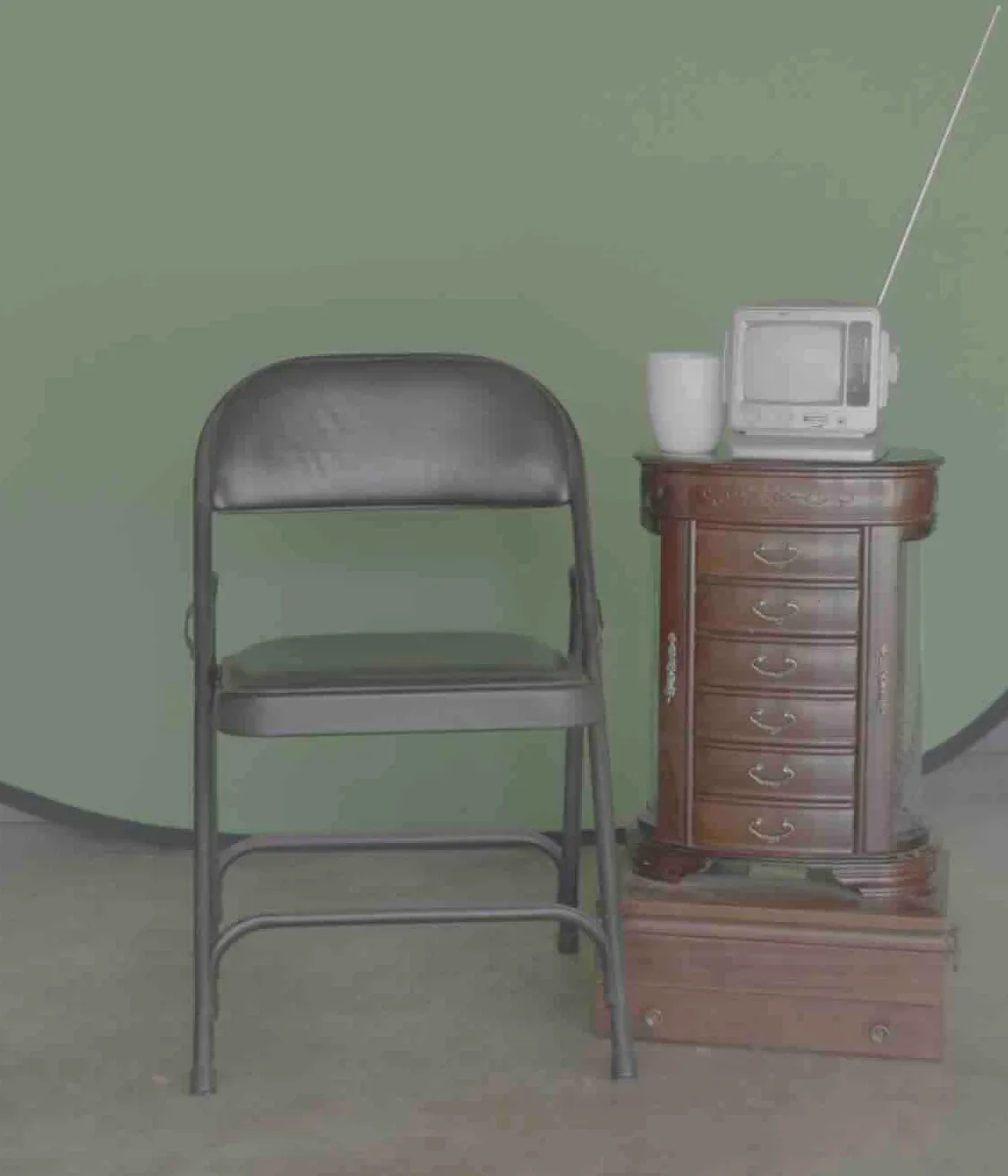 A folding chair stands next to an antique column of drawers. A tv and a coffee mug sit on top of the drawers.